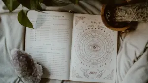 Atversta knyga su zodiako ženklais ant jaukaus užkloto šalia kristalo.